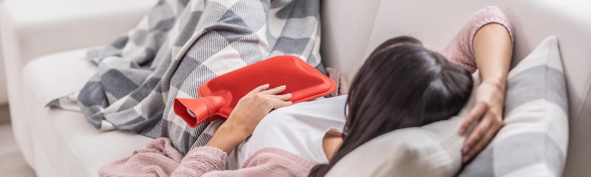 Eine Frau liegt mit Menstruationsbeschwerden auf dem Sofa. Sie hält eine rote Wärmflasche auf ihren Bauch und ist mit einer karierten Decke zugedeckt.