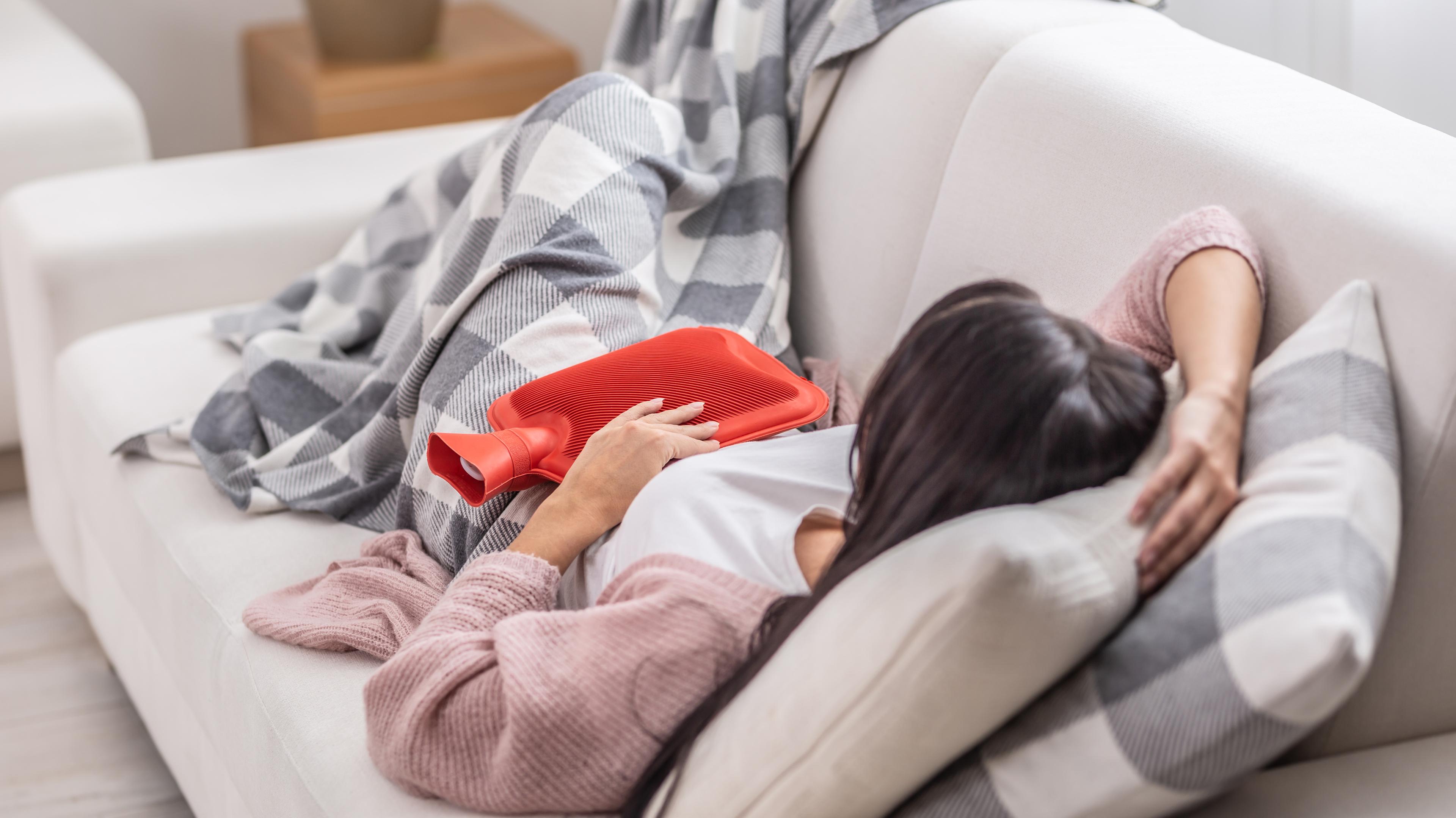 Eine Frau liegt mit Menstruationsbeschwerden auf dem Sofa. Sie hält eine rote Wärmflasche auf ihren Bauch und ist mit einer karierten Decke zugedeckt.