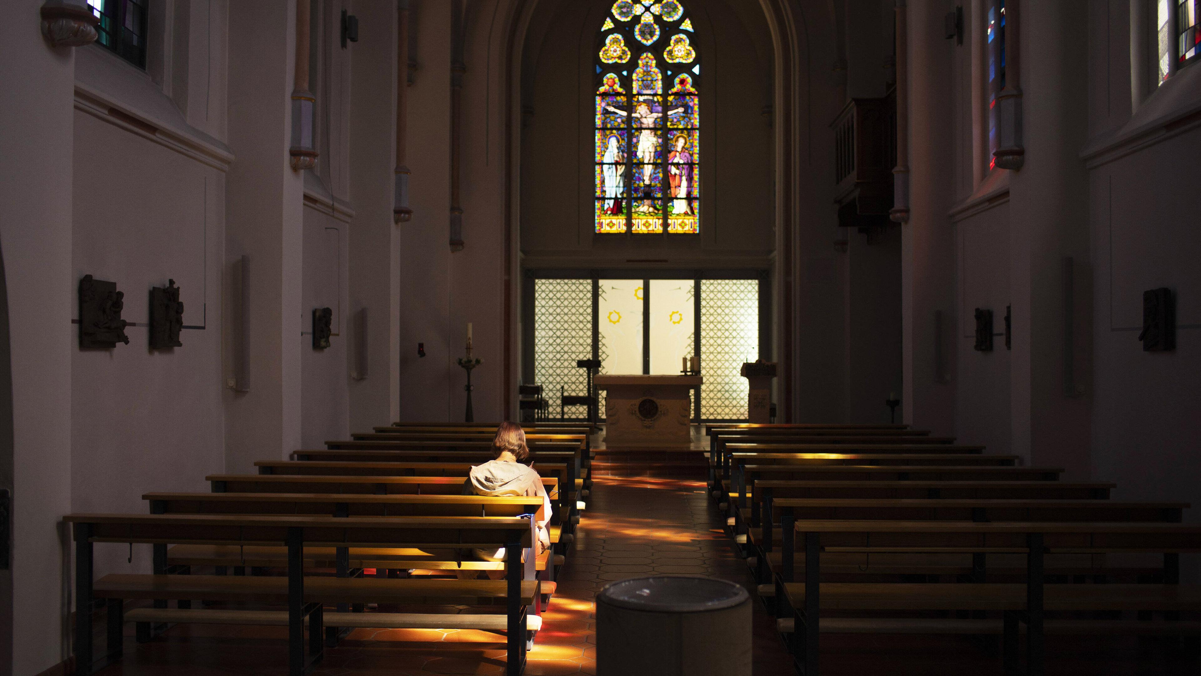 Rückansicht einer Frau, die alleine in einer Kirchenbank in einer sonst leeren alten Kirche mit einem großen Buntglasfenster über dem Altar sitzt.