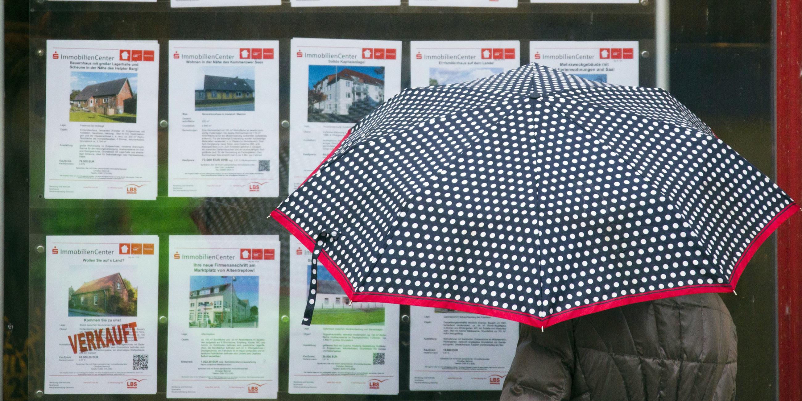 Archiv: Eine Frau mit Regenschirm steht vor einem Schaufenster mit Ammobilienangeboten.