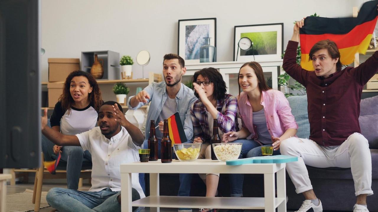 Typical Fußballzuschauer auf Couch: Gruppe junger Leute mit Deutschlandfahnen sitzt angespannt auf Sofa und schaut Richtung Fernseher