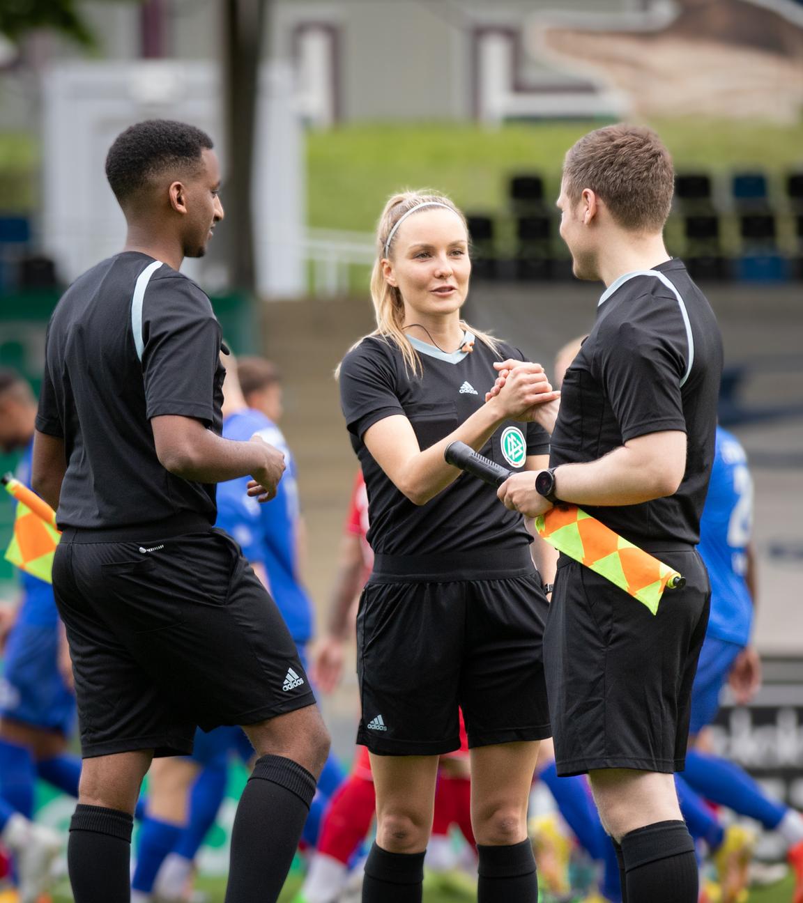 Fabienne ist im Stadion in Schiedsrichteroutfit im Gespräch mit zwei männlichen Kollegen. Im Hintergrund laufen Fußballspieler in blauer oder roter Trainingskleidung.
