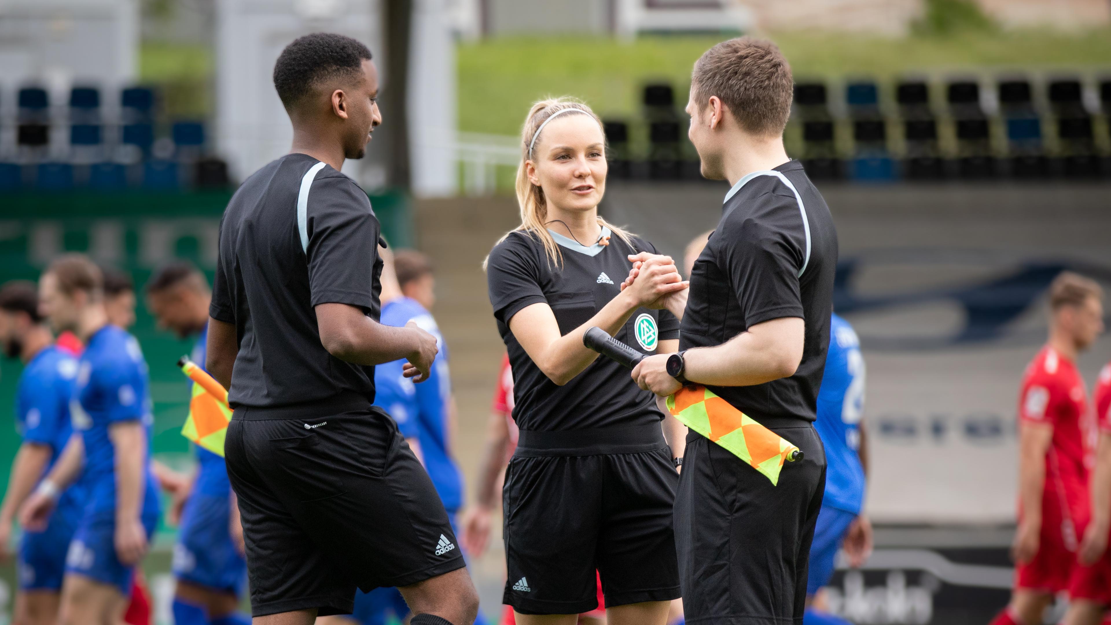 Fabienne ist im Stadion in Schiedsrichteroutfit im Gespräch mit zwei männlichen Kollegen. Im Hintergrund laufen Fußballspieler in blauer oder roter Trainingskleidung.