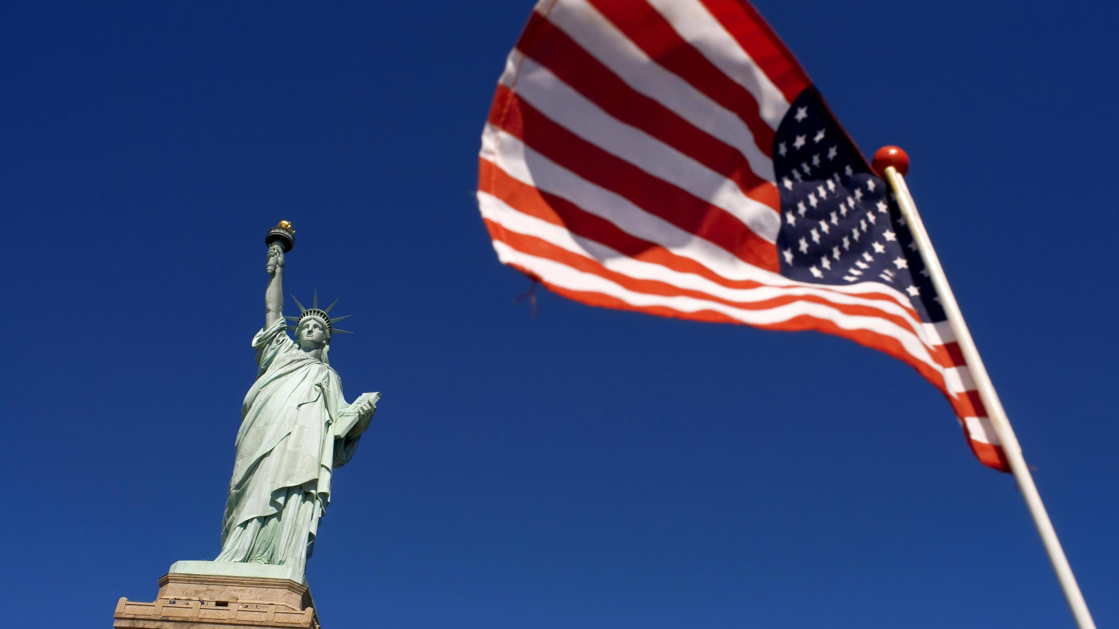 Untersicht auf eine Freiheitsstatue - eine Frau mit Sternenkrone, die eine Fackel hochhält. Daneben flattert eine US-Flagge vor blauem Himmel.