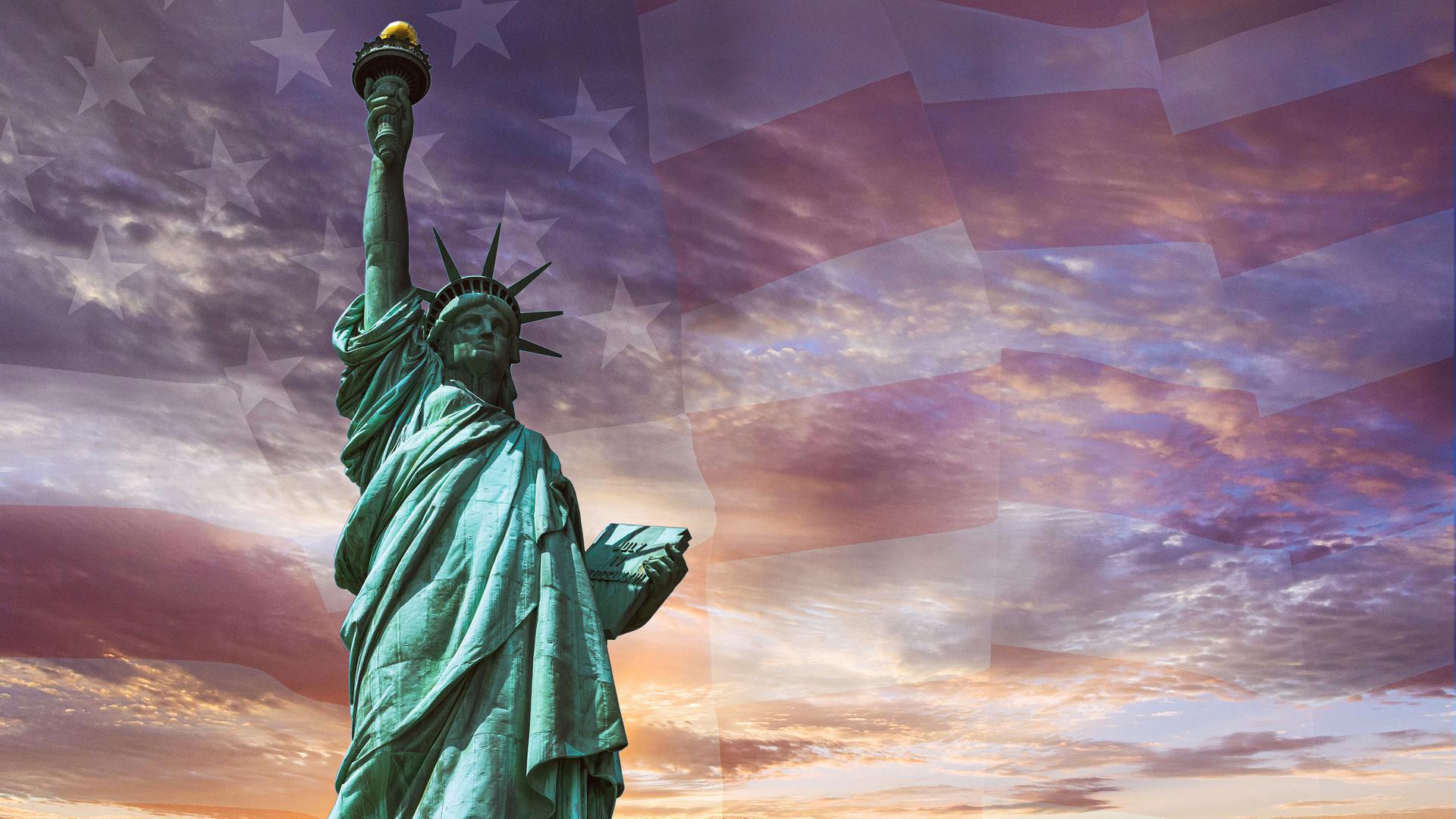 Die Freiheitsstatue - eine Frau mit Sternenkrone hält eine Fackel hoch - vor verschwommener US-Flagge