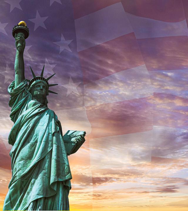 Die Freiheitsstatue - eine Frau mit Sternenkrone hält eine Fackel hoch - vor verschwommener US-Flagge