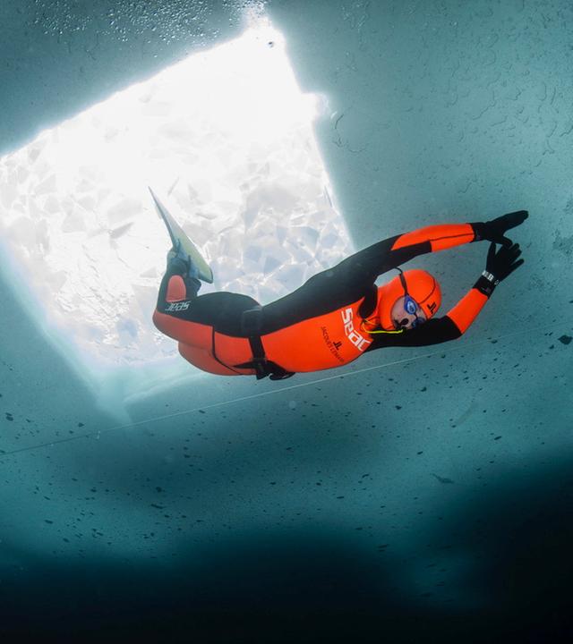 Freitaucherin Valentina Cafolla taucht in einem scharz-orangenen Neoprenanzug unter dem Eis. Über ihr sieht man ein viereckiges Loch im Eis. 
