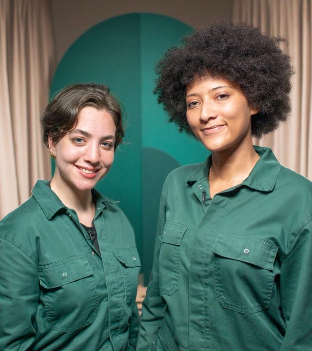 Zwei Frauen in grünen Overalls blicken in die Kamera