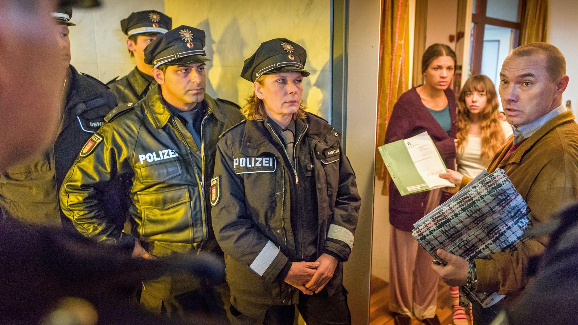 "Notruf Hafenkante - Fremde Heimat": Vor einer geöffneten Wohnungstür steht Bernd Dillinger (Thomas Arnold). Er hält einen Beschluss in der rechten Hand und sieht zu einem Polizisten (Komparse), der hinter ihm steht. Weitere vier Beamte, darunter Tarik Coban (Serhat Cokgezen) und Claudia Fischer (Janette Rauch), (Rest Komparsen) stehen hintereinander seitlich der Wohnungstür und schauen Dillinger an. In der Tür stehen Eva (Isabelle Redfern) und Isabella Escobar (Lisa Brand), die ängstlich zu Dillinger sehen.