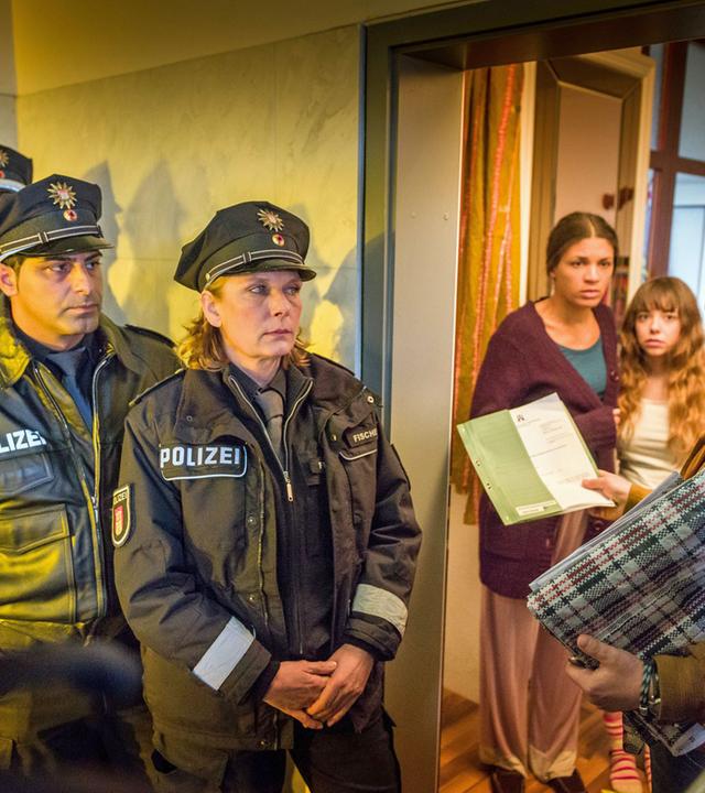 "Notruf Hafenkante - Fremde Heimat": Vor einer geöffneten Wohnungstür steht Bernd Dillinger (Thomas Arnold). Er hält einen Beschluss in der rechten Hand und sieht zu einem Polizisten (Komparse), der hinter ihm steht. Weitere vier Beamte, darunter Tarik Coban (Serhat Cokgezen) und Claudia Fischer (Janette Rauch), (Rest Komparsen) stehen hintereinander seitlich der Wohnungstür und schauen Dillinger an. In der Tür stehen Eva (Isabelle Redfern) und Isabella Escobar (Lisa Brand), die ängstlich zu Dillinger sehen.