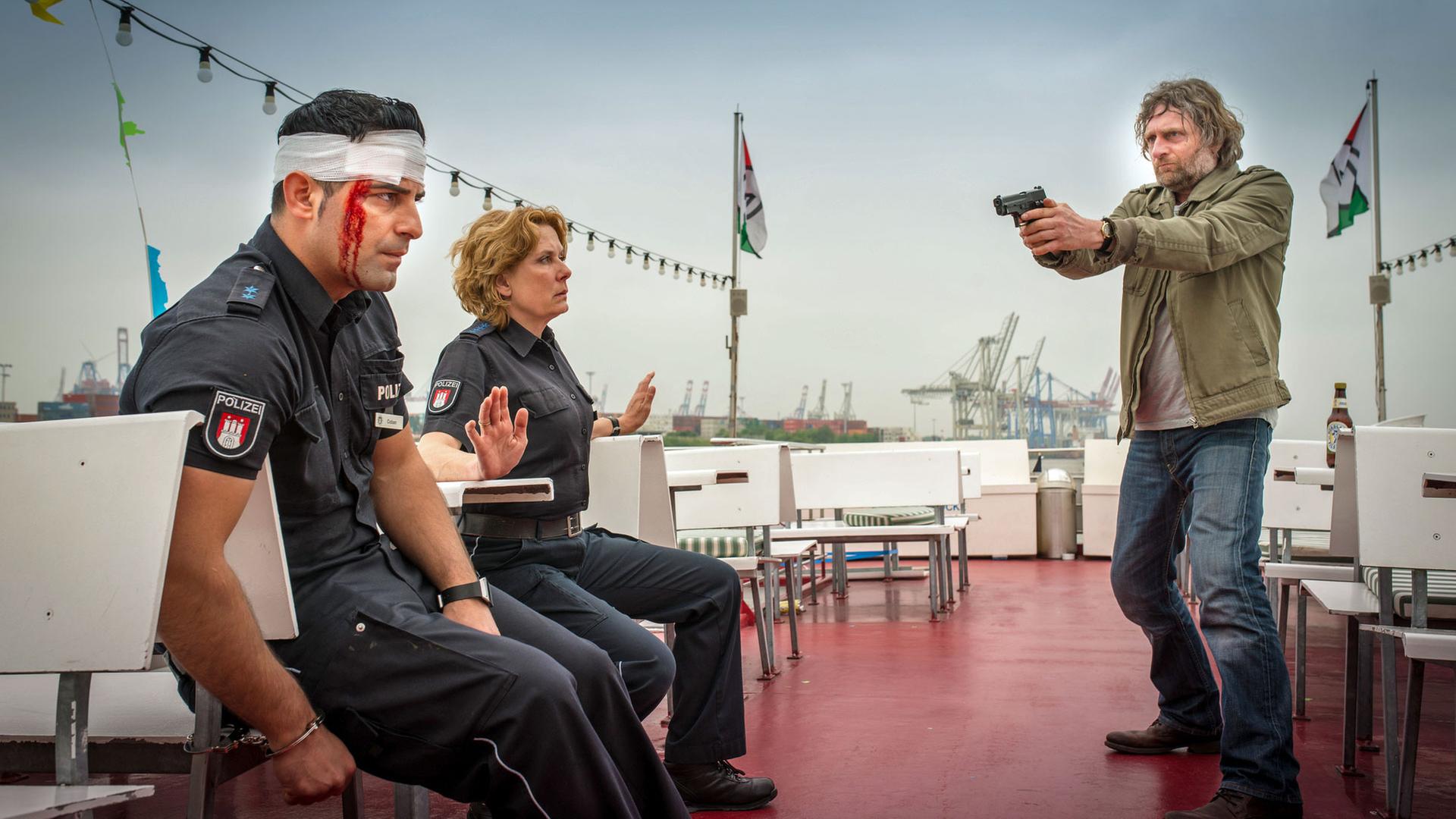 "Notruf Hafenkante - Fremder Vater": Malte Lampert (Jörg Witte) steht auf einem Ausflugsboot. Er hält eine Waffe mit beiden Händen und zielt damit auf die Polizisten Tarik Coban (Serhat Cokgezen) und Claudia Fischer (Janette Rauch). Sie sitzen ihm gegenüber auf Holzbänken. Tarik ist mit einer Handschelle an eine Bank gefesselt, Claudia hebt die Hände. Tarik hat einen Verband um den Kopf, Blut läuft von seiner rechten Schläfe herunter.
