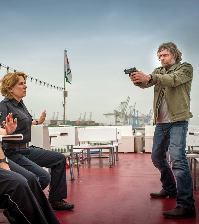 "Notruf Hafenkante - Fremder Vater": Malte Lampert (Jörg Witte) steht auf einem Ausflugsboot. Er hält eine Waffe mit beiden Händen und zielt damit auf die Polizisten Tarik Coban (Serhat Cokgezen) und Claudia Fischer (Janette Rauch). Sie sitzen ihm gegenüber auf Holzbänken. Tarik ist mit einer Handschelle an eine Bank gefesselt, Claudia hebt die Hände. Tarik hat einen Verband um den Kopf, Blut läuft von seiner rechten Schläfe herunter.