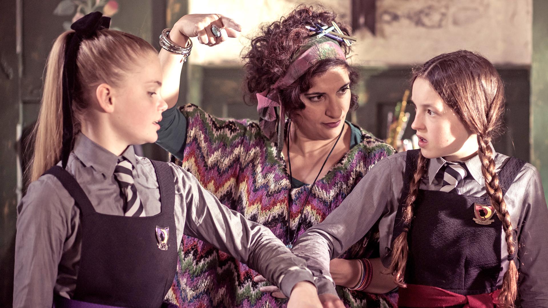 Frau Schimmel zeigt auf die Freundschaftsfalle, die Esther und Mildred an den Handgelenken miteinander verknüpft. Mildred ist nicht begeistert davon.