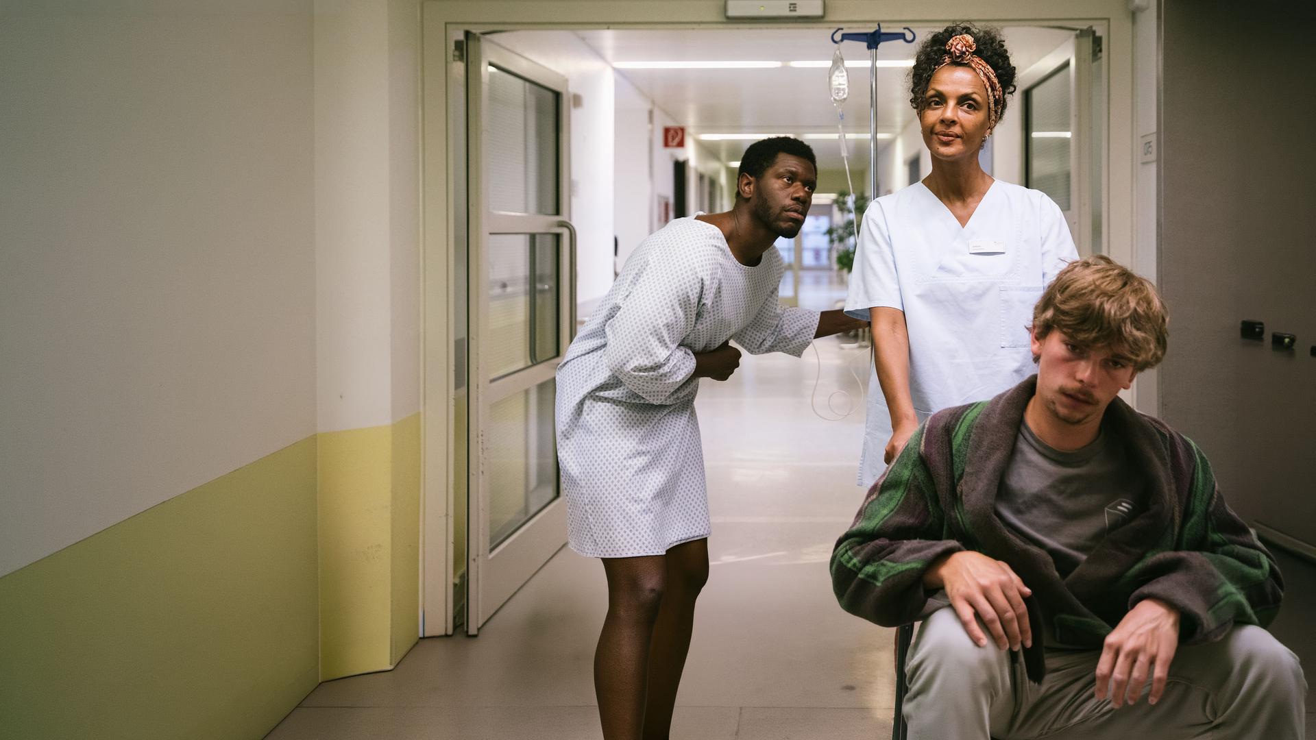 "I don't work here - Friedhof": Dawit (Akeem van Flodrop) steht gebückt in einem Krankenhausflur und hält sich mit der rechten Hand den Bauch. Er trägt ein weißes Krankenhaushemd und hält sich mit links an einem Infusionsständer fest. Er schaut seiner Mutter Lemlem (Dennenesch Zoudé) hinterher, die in ihrer Pflegerinnenkleidung einen Patienten im Rollstuhl vor sich herschiebt und dabei lächelt.