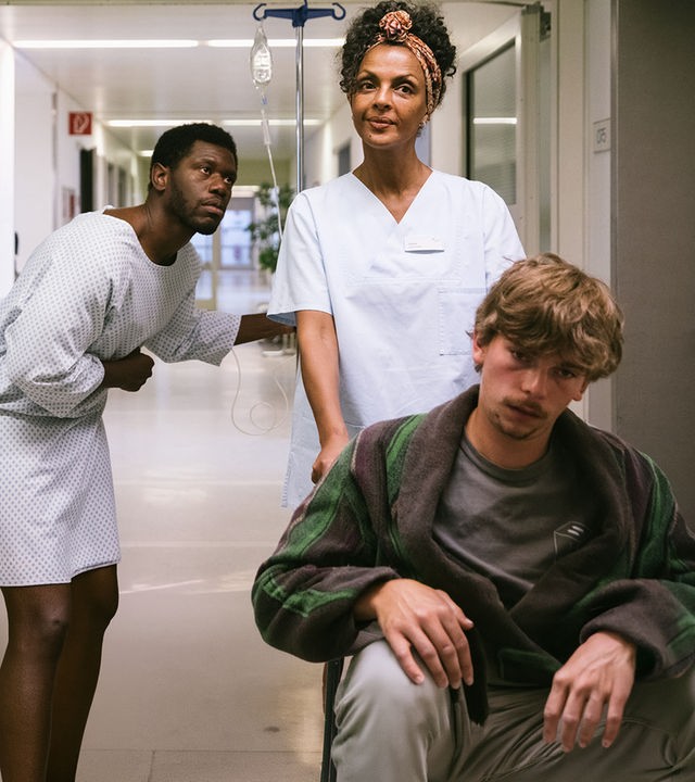 "I don't work here - Friedhof": Dawit (Akeem van Flodrop) steht gebückt in einem Krankenhausflur und hält sich mit der rechten Hand den Bauch. Er trägt ein weißes Krankenhaushemd und hält sich mit links an einem Infusionsständer fest. Er schaut seiner Mutter Lemlem (Dennenesch Zoudé) hinterher, die in ihrer Pflegerinnenkleidung einen Patienten im Rollstuhl vor sich herschiebt und dabei lächelt.