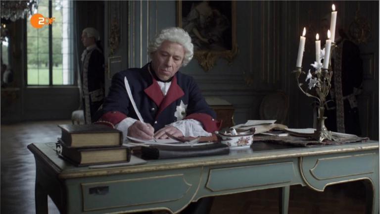 Friedrich der Große sitzt schreibend an seinem Schreibtisch.