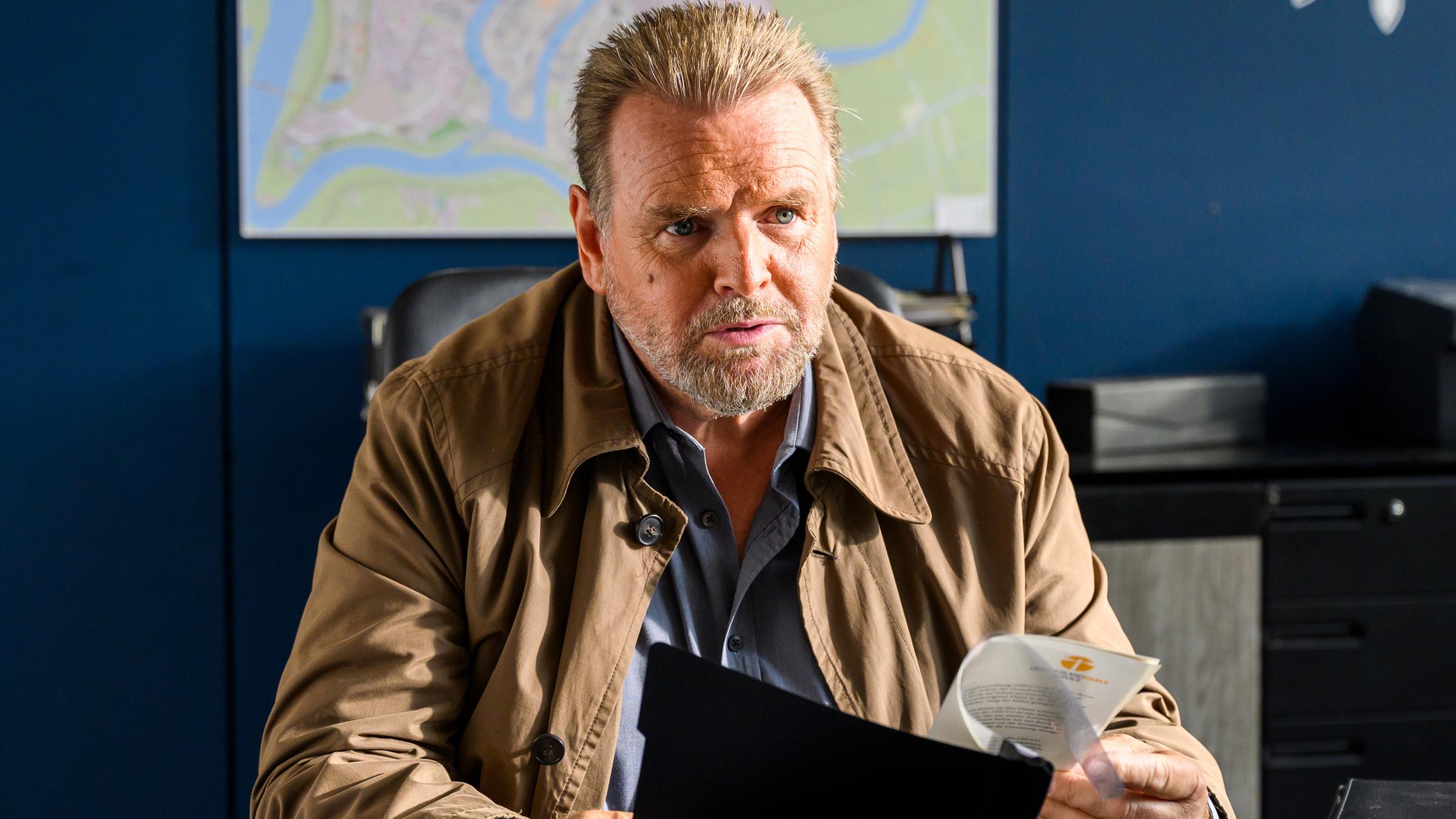 Brockhorst (Felix Vörtler) sitzt an seinem Schreibtisch vor einer blauen Wand auf der eine Landkarte hängt und hält einen Schnellhefter in den Händen.