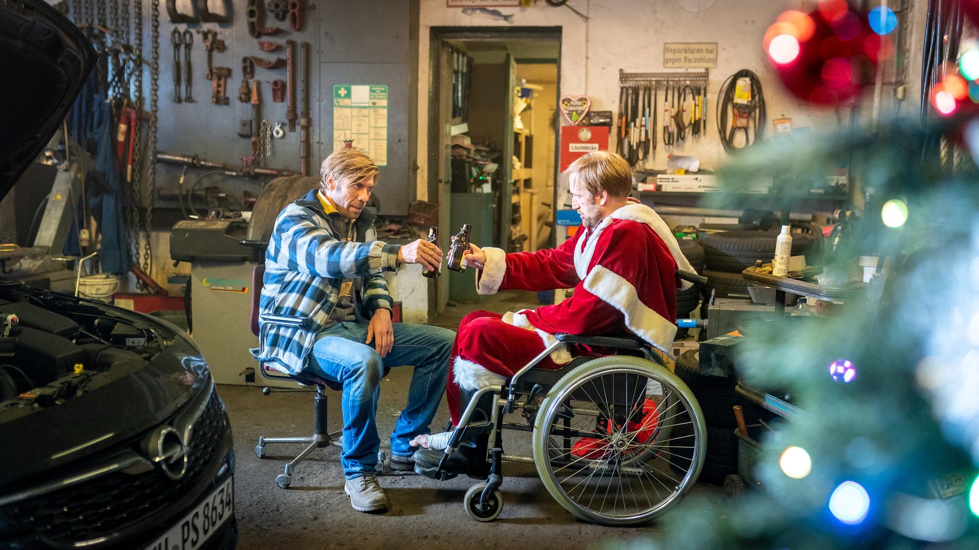 "Notruf Hafenkante - Frohe Weihnachten": Hannes (Maik Rogge) sitzt in einem Rollstuhl. Er trägt ein Weihnachtsmannkostüm. Ihm gegenüber sitzt Lutz (Felix Lampert). Beide prosten sich mit einer Bierflasche zu. Sie befinden sich in einer Werkstatt.