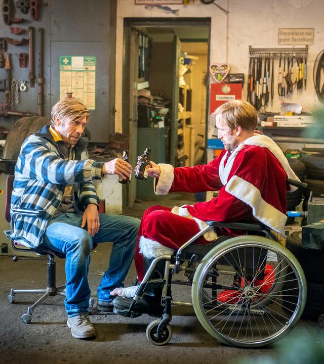 "Notruf Hafenkante - Frohe Weihnachten": Hannes (Maik Rogge) sitzt in einem Rollstuhl. Er trägt ein Weihnachtsmannkostüm. Ihm gegenüber sitzt Lutz (Felix Lampert). Beide prosten sich mit einer Bierflasche zu. Sie befinden sich in einer Werkstatt.