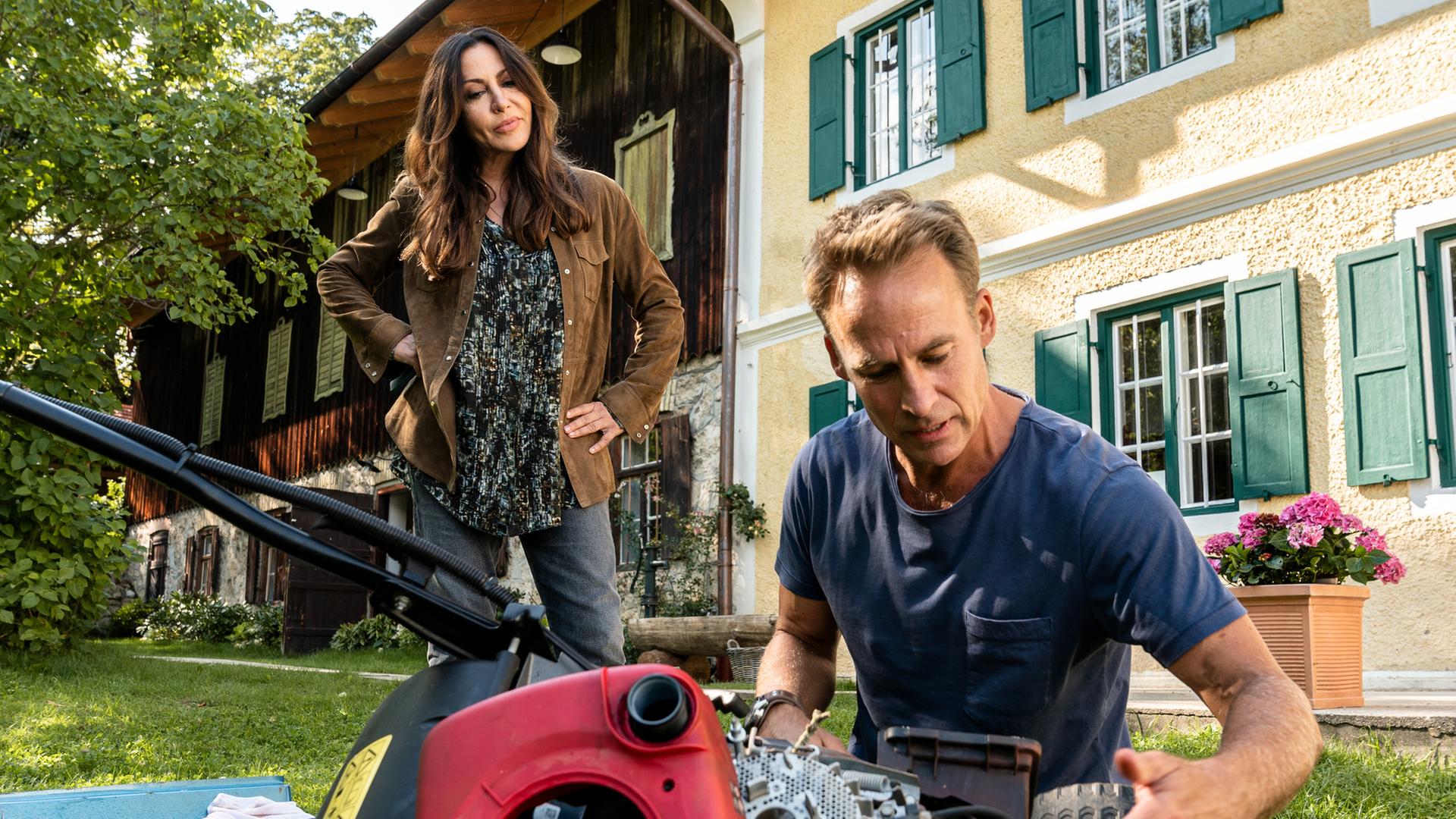 "Frühling - Alte Gespenster": Katja (Simone Thomalla) schaut stehend zu, wie Mark (Marco Girnth) den Rasenmäher repariert. Im Hintergrund das gelbe Haus mit grünen Fensterläden, in dem Katja wohnt.