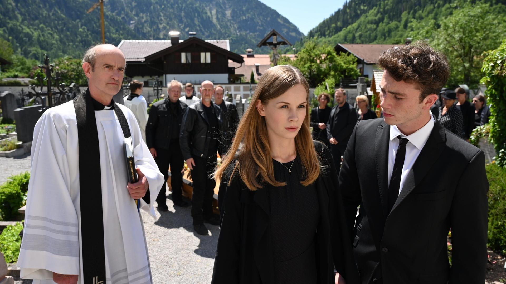 "Frühling - Genieße jeden Augenblick": Johanna Brugger (Emilia Bernsdorf) und Gerry (Justus Johanssen) gehen nebeneinander her. Beide tragen Trauerkleidung. Pfarrer Sonnleitner (Johannes Herrschmann) blickt ihnen nach.