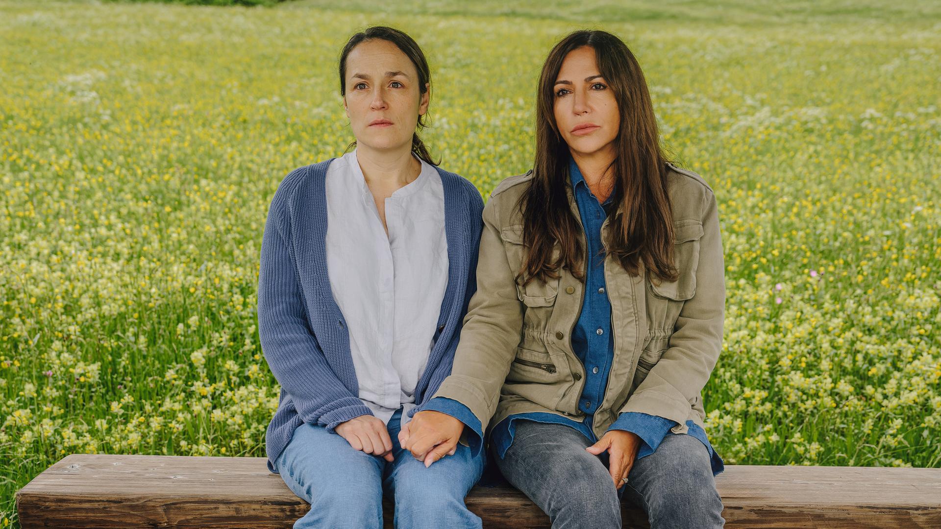 Katja Baumann (Simone Thomalla) sitzt mit Maria Ronzheimer (Judith Toth) auf einer Bank auf einer Wiese und hält ihre Hand.