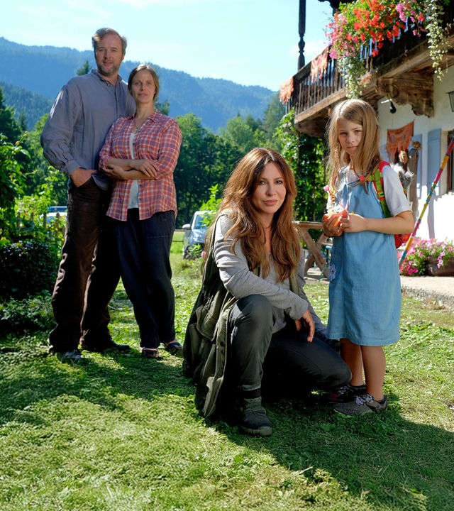 "Frühling - Wenn Kraniche fliegen": Lea Krug (Elin Paasch) steht vor einem bayerischen Hof mit bunten Blumenkästen. Neben ihr kniet Katja Baumann (Simone Thomalla). Im Hintergrund stehen Gregor Krug (Stephan Grossmann) und Stefanie Krug (Julia Jäger). Im Hintergrund bewaldete Berge und blauer Himmel.