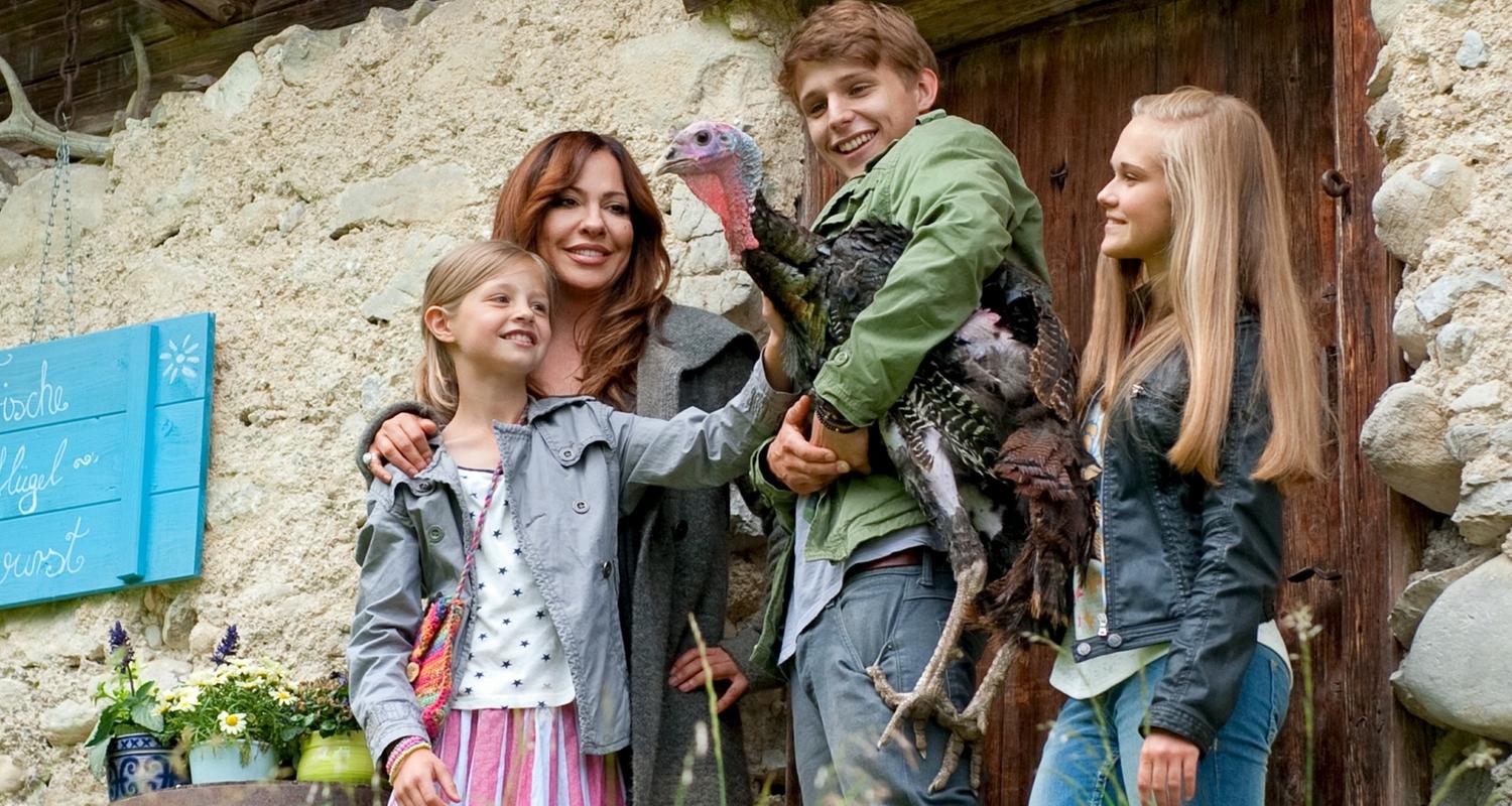 Katja mit den drei Kindern auf dem elternlosen Hof der Familie Böhm. Der Sohn hält einen Truthahn im Arm, der von allen beobachtet wird (von links: Flora Li Thiemann, Simone Thomalla, Merlin Rose, Nadine Kösters).