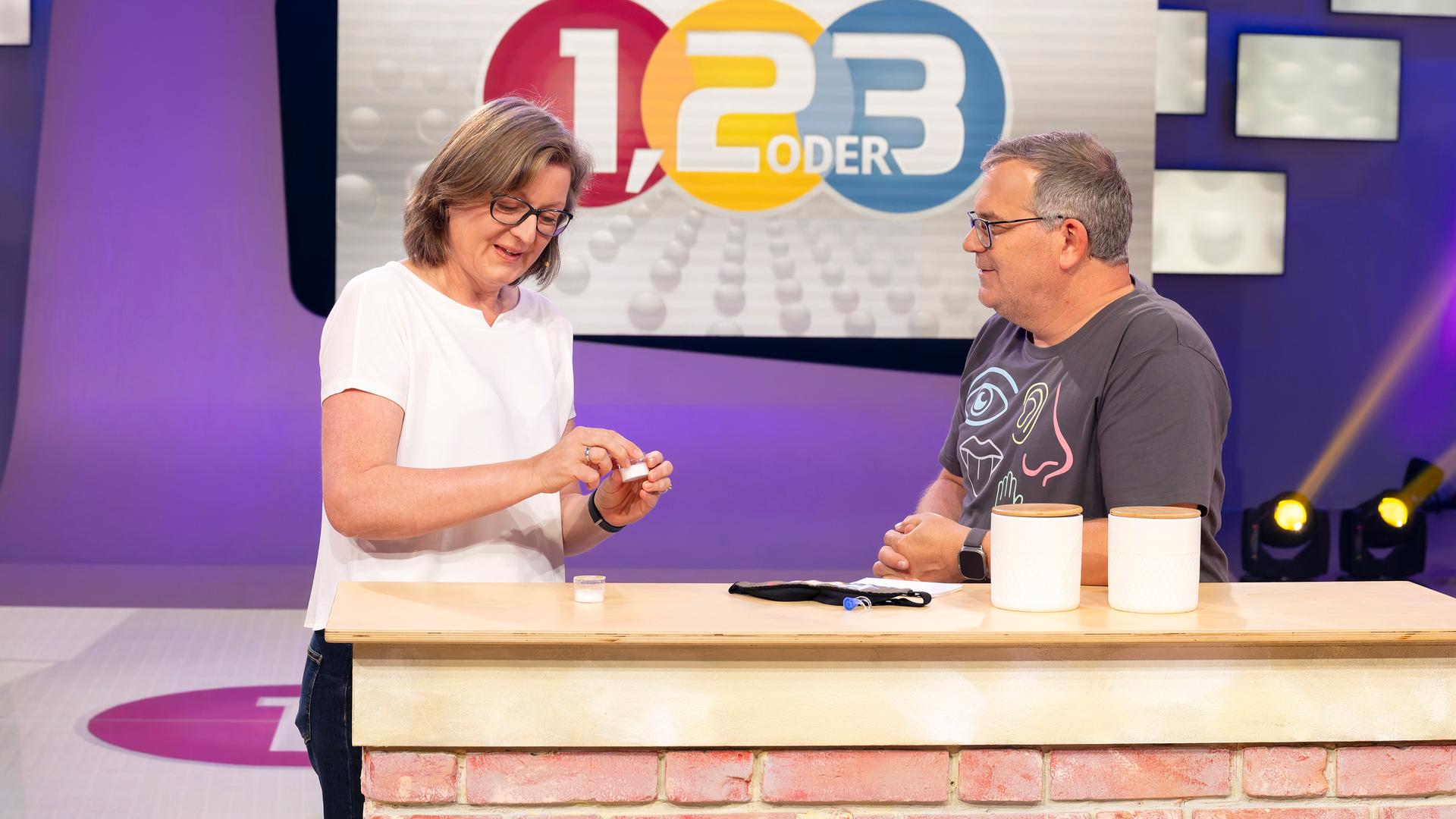 "1, 2 oder 3 - Fühlen, riechen, staunen": Prof. Dr. Jessica Freiherr steht mit Elton hinter einem Pult im Studio. Sie hält eine Duftprobe in einem kleinen Glasbehälter in der Hand. Im Hintergrund ist das "1, 2 oder 3"- Logo zu sehen.