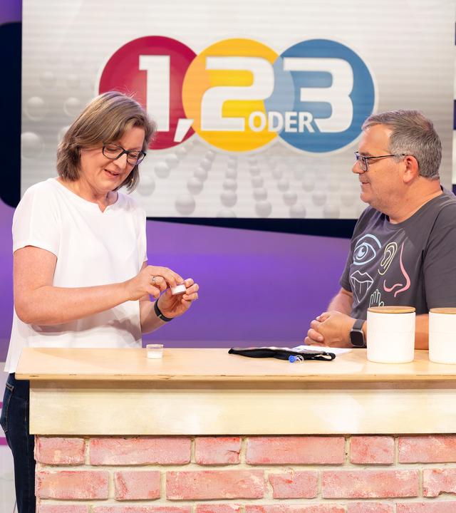 "1, 2 oder 3 - Fühlen, riechen, staunen": Prof. Dr. Jessica Freiherr steht mit Elton hinter einem Pult im Studio. Sie hält eine Duftprobe in einem kleinen Glasbehälter in der Hand. Im Hintergrund ist das "1, 2 oder 3"- Logo zu sehen.