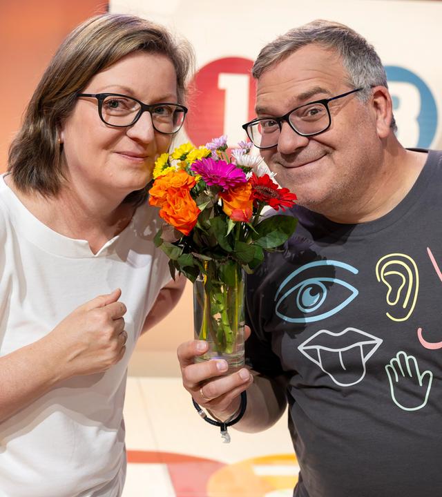 "1, 2 oder 3 - Fühlen, riechen, staunen": Prof. Dr. Jessica Freiherr steht mit Elton hinter einem Pult im Studio. Sie hält eine Duftprobe in einem kleinen Glasbehälter in der Hand. Im Hintergrund ist das "1, 2 oder 3"- Logo zu sehen.