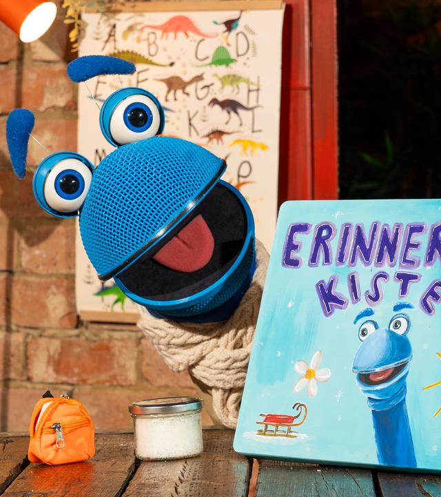 Das SingDing schaut mit weit geöffnetem Mund hinter seiner hellblauen Erinnerungskiste hervor, die auf dem Tisch steht. Es trägt einen beigen Schal. Vor ihm auf dem Tisch stehen ein kleiner oranger Rucksack und ein Glas voll Schnee.