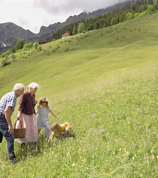 Im Bild sind drei Personen und ein Kind in einer grünen Wiese zu sehen. Sie gehen in Richtung einer Berglandschaft mit bewaldeten Hängen. Eine Person hält einen Korb.
