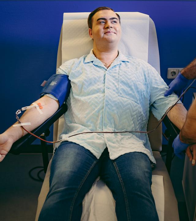 Nico liegt auf einem Liegestuhl bei Plasmaspende. Er hat seinen Arm ausgestreckt, am Arm ist eine Manschette befestigt, darunter befindet sich ein Gerät mit einem Schlauch. Neben ihm, im Anschnitt zu erkennen, steht eine Pflegekraft.