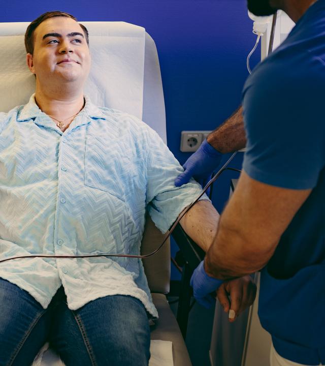 Nico liegt auf einem Liegestuhl bei der Plasmaspende. Er hat seinen Arm ausgestreckt, am Arm ist eine Manschette befestigt, darunter befindet sich ein Gerät mit einem Schlauch. Neben ihm, im Anschnitt zu erkennen, steht eine Pflegekraft.