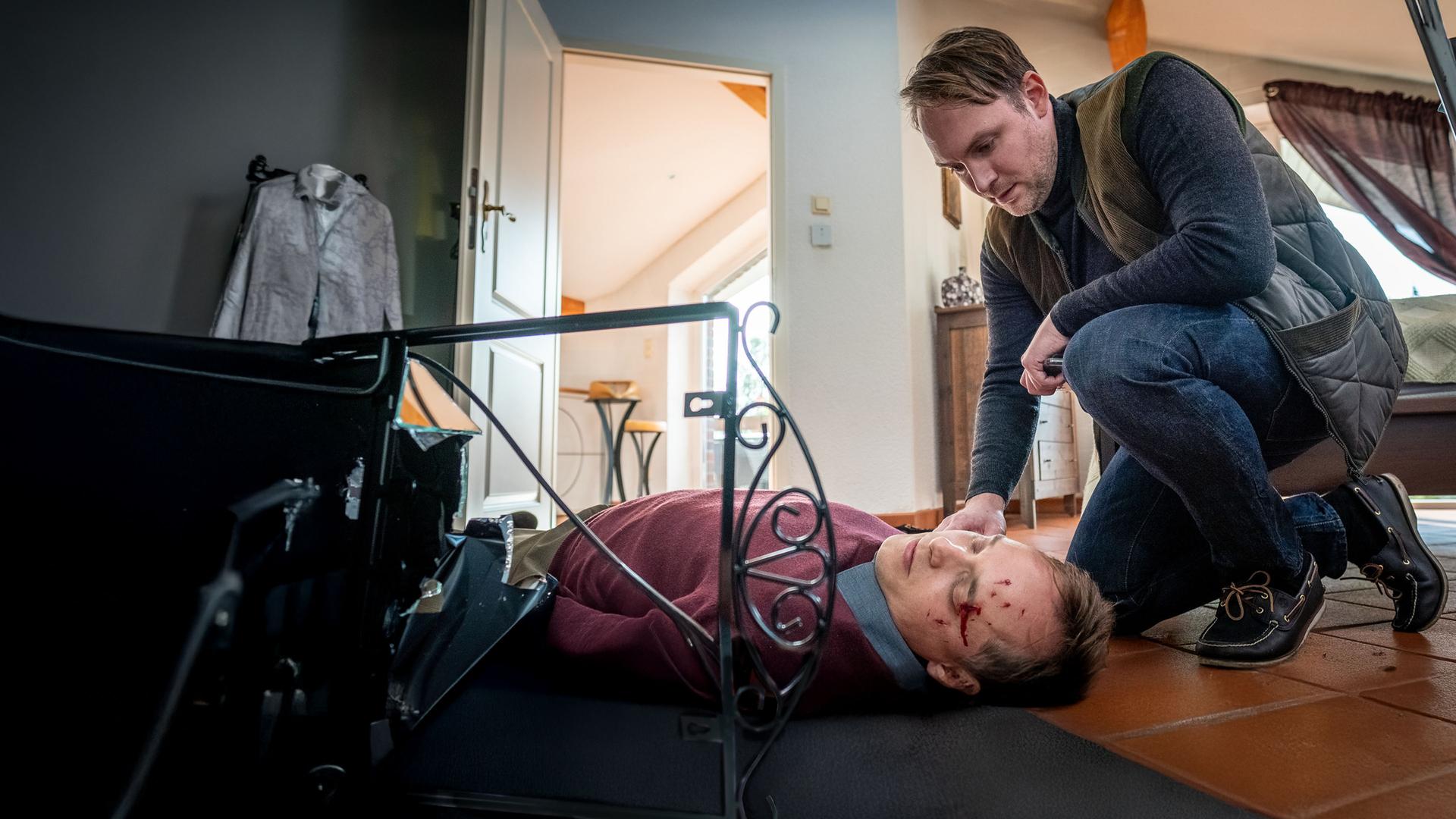 "Notruf Hafenkante - Für eine Handvoll Euro": Justin Hertels (Adrian Zwicker) liegt bewusstlos auf dem Rücken auf dem Fußboden in seinem Haus. Thies Scholtens (Martin Stange) kniet in der Hocke neben ihm.