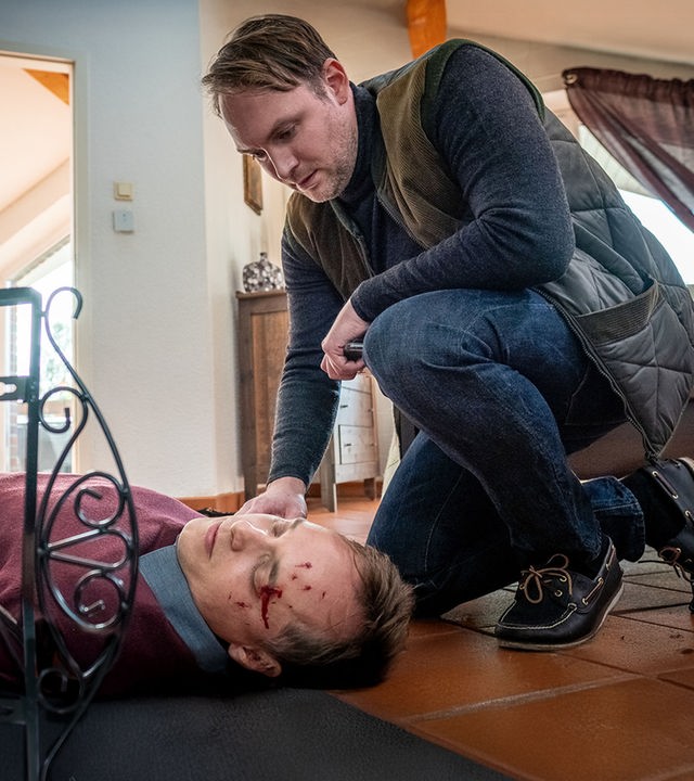 "Notruf Hafenkante - Für eine Handvoll Euro": Justin Hertels (Adrian Zwicker) liegt bewusstlos auf dem Rücken auf dem Fußboden in seinem Haus. Thies Scholtens (Martin Stange) kniet in der Hocke neben ihm.