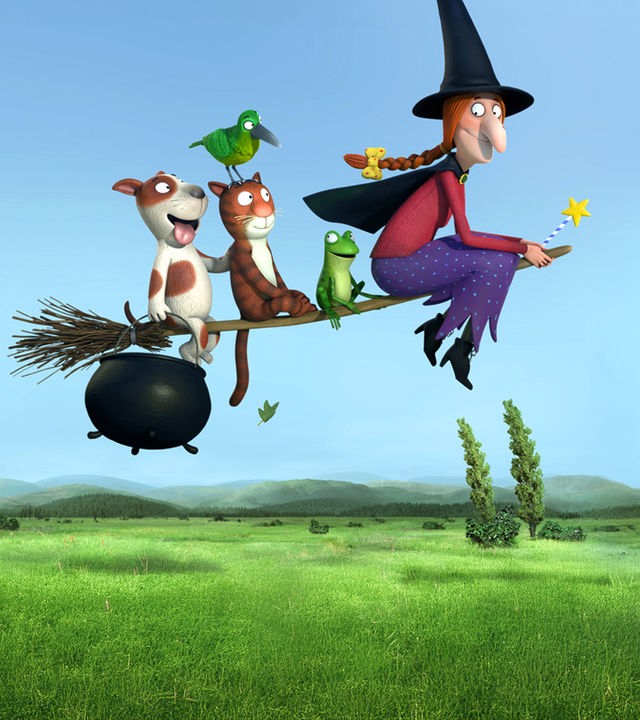 Eine Hexe und ihre Katze sitzen auf einem Hexenbesen. Die Hexe hält mit der rechten Hand einen Hexenkessel und mit der linken Hand ihren Zauberstab. Sie fliegen durch die Luft. Unter ihnen sieht man Berge, Felder und Wälder.. 