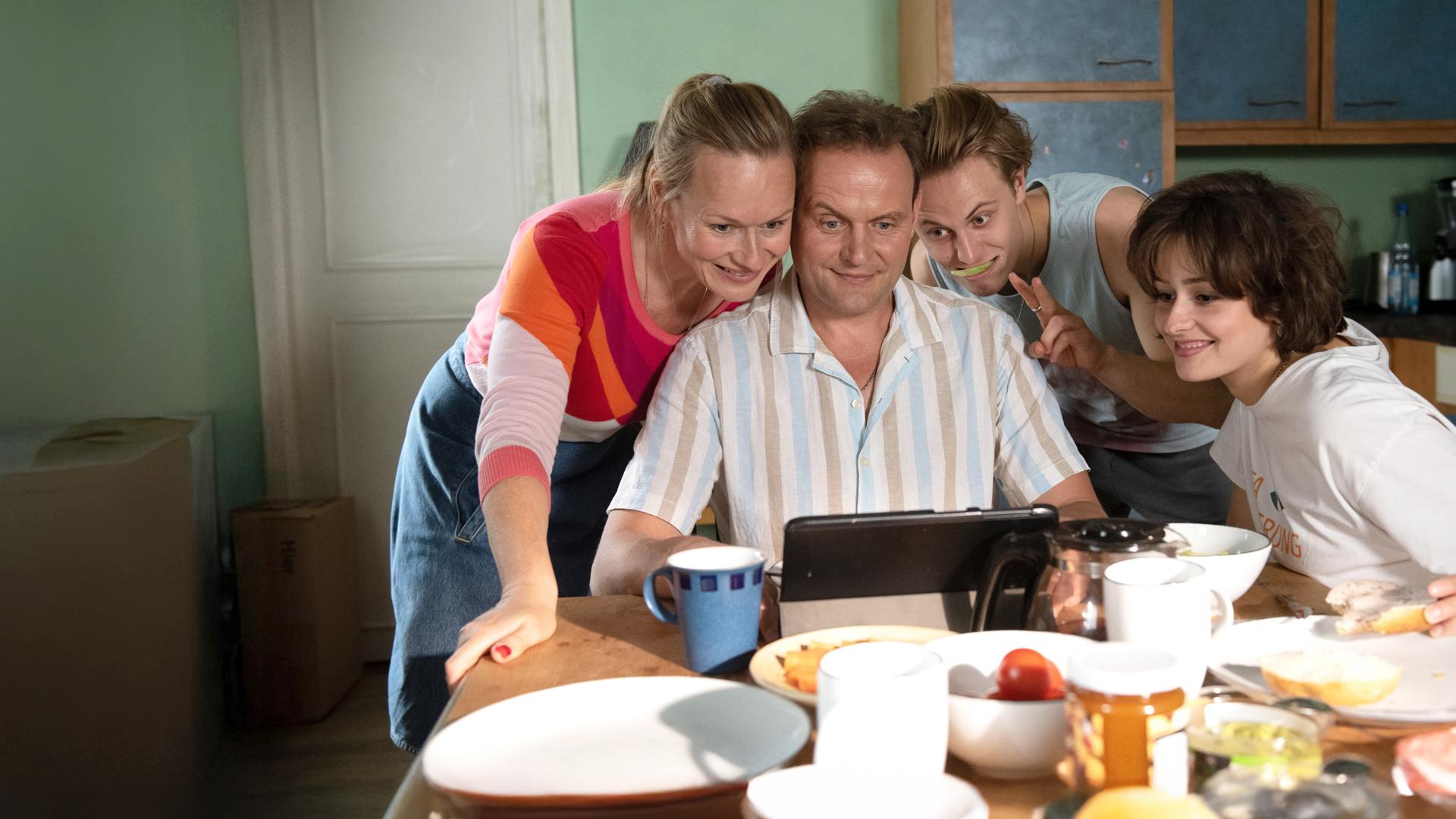"Für immer Eltern": Michael Wagner (Devid Striesow) sitzt an einem gedeckten Frühstückstisch und schaut lächelnd auf ein Tablet, das er in der Hand hält. Seine Frau Anja (Anja Schneider), steht links neben ihm und schaut ebenfalls auf das Tablet, ebenso wie ihr Sohn Niklas (Max Schimmelpfennig), rechts neben Michael und dessen Freundin Alina (Anouk Elias) ganz rechts am Tisch.