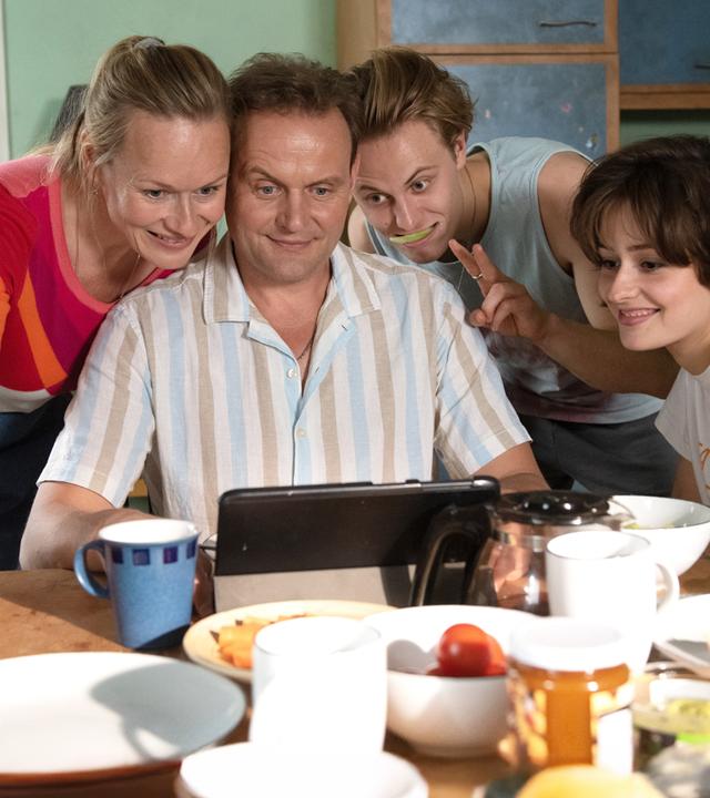 "Für immer Eltern": Michael Wagner (Devid Striesow) sitzt an einem gedeckten Frühstückstisch und schaut lächelnd auf ein Tablet, das er in der Hand hält. Seine Frau Anja (Anja Schneider), steht links neben ihm und schaut ebenfalls auf das Tablet, ebenso wie ihr Sohn Niklas (Max Schimmelpfennig), rechts neben Michael und dessen Freundin Alina (Anouk Elias) ganz rechts am Tisch.