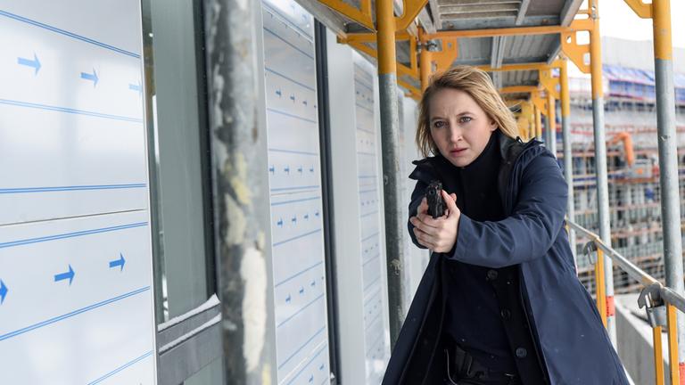 Nora (Anna Maria Mühe) steht mit gezogener Waffe auf einem Baugerüst einer Baustelle.