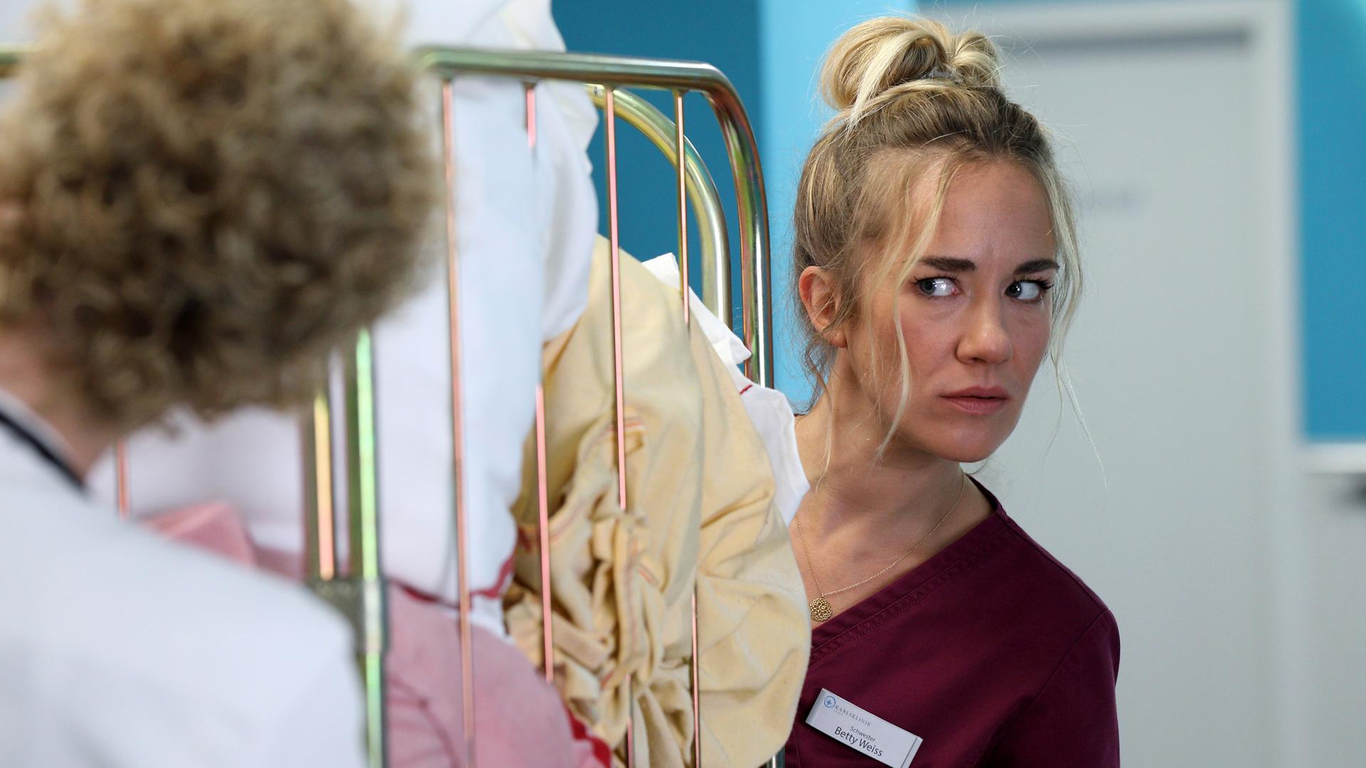 Dr. Helena von Arnstett (Claudia Hiersche) steht auf dem Krankenhausflur vor einem Wäschewagen. Betty (Annina Hellenthal) steht hinter dem Wagen und schaut sie skeptisch an.