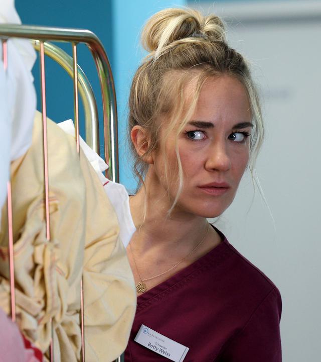 Dr. Helena von Arnstett (Claudia Hiersche) steht auf dem Krankenhausflur vor einem Wäschewagen. Betty (Annina Hellenthal) steht hinter dem Wagen und schaut sie skeptisch an.