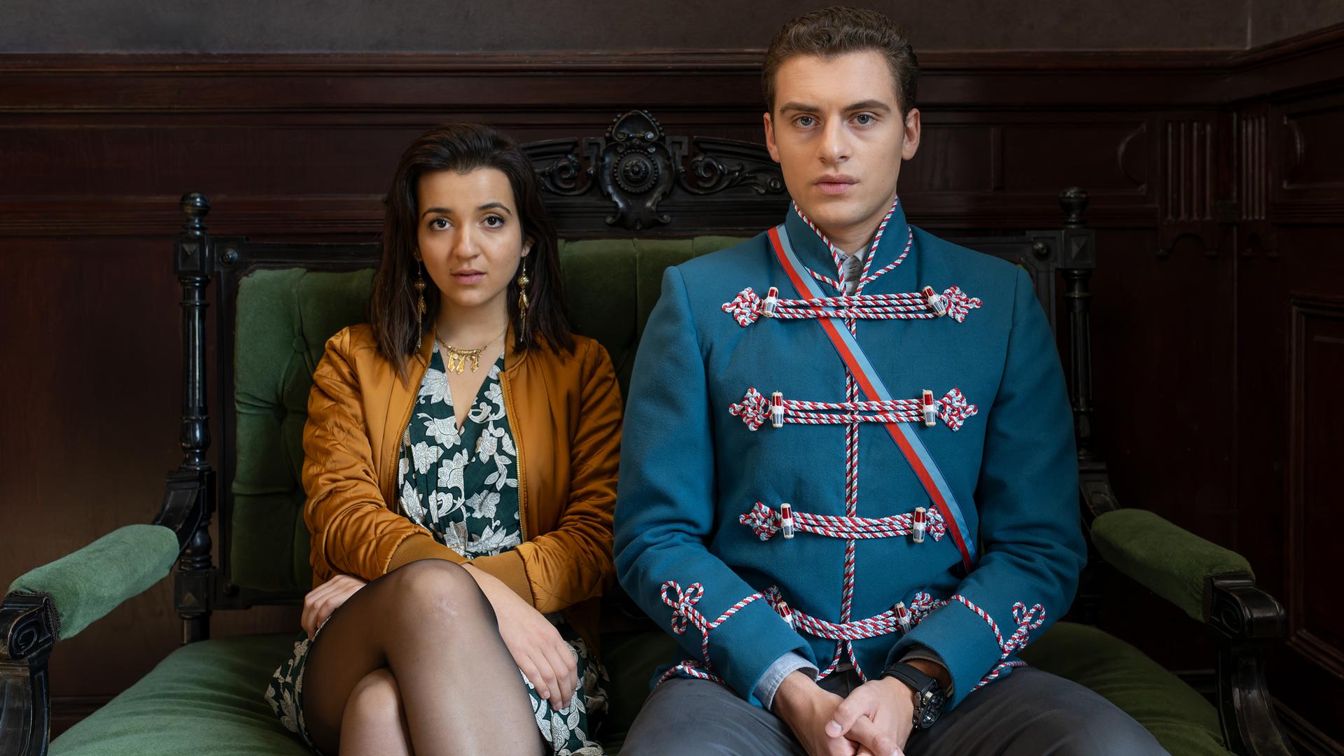 "Füxe": Mina (Roxana Samadi) und Adem (Valon Krasniqi) sitzen nebeneinander in aufrechter Haltung auf einem antiken Sofa, das mit grünem Samt bespannt ist.