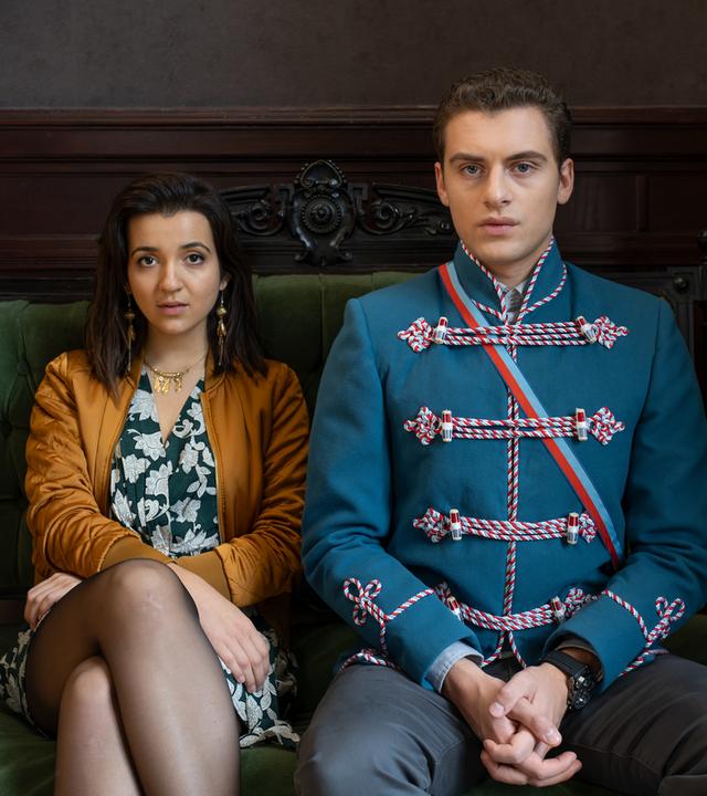 "Füxe": Mina (Roxana Samadi) und Adem (Valon Krasniqi) sitzen nebeneinander in aufrechter Haltung auf einem antiken Sofa, das mit grünem Samt bespannt ist.