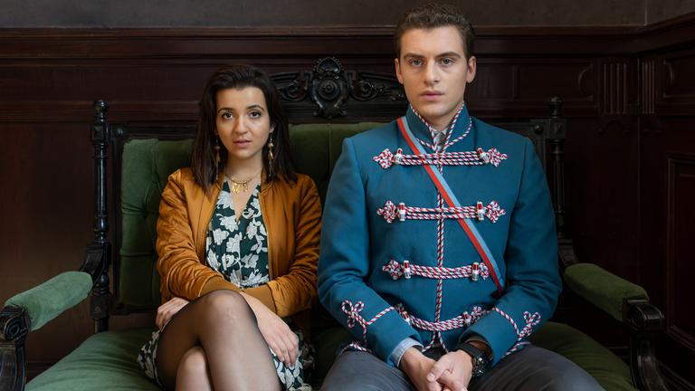 "Füxe": Mina (Roxana Samadi) und Adem (Valon Krasniqi) sitzen nebeneinander in aufrechter Haltung auf einem antiken Sofa, das mit grünem Samt bespannt ist.