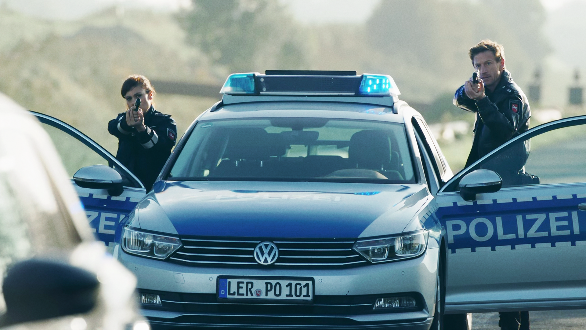 Süher Özlügül (Sophia Dal) und Henk Cassens (Maxim Mehmet) stehen mit ehobenen Waffen am Polizeiauto. 