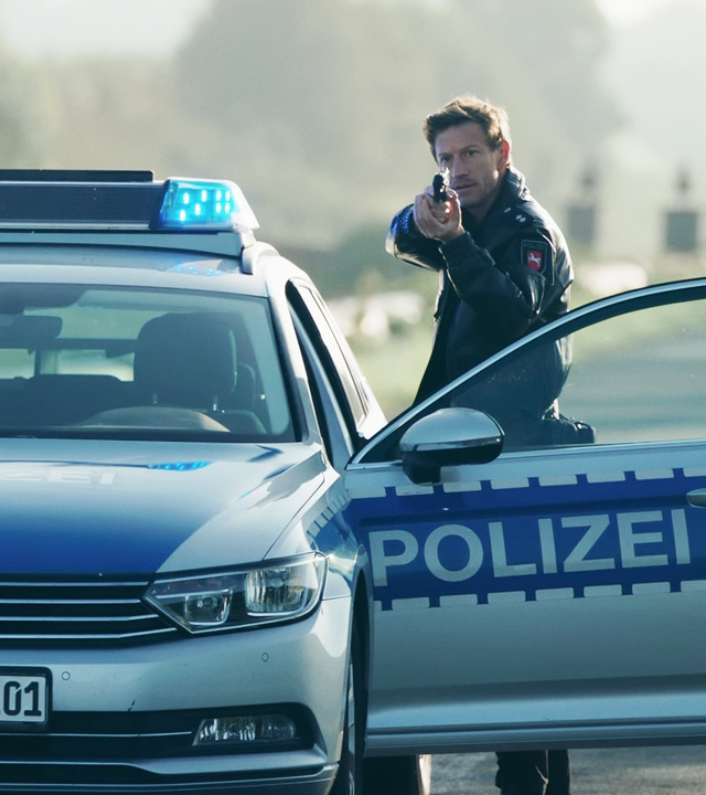 Süher Özlügül (Sophia Dal) und Henk Cassens (Maxim Mehmet) stehen mit ehobenen Waffen am Polizeiauto. 