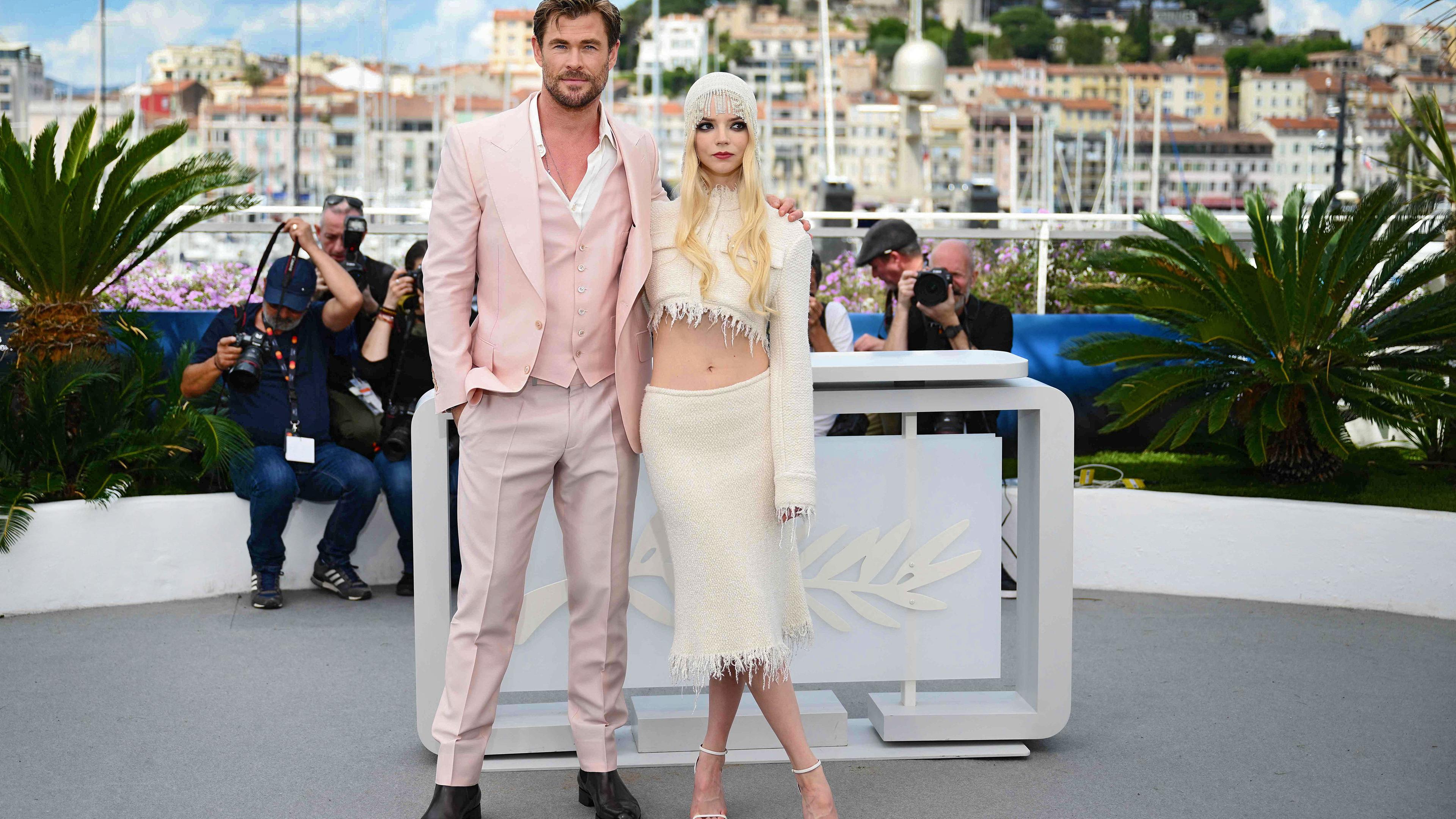 Ein Mann im lässig offenen rosa Jackett mit Weste und eine Frau in einem bauchfreien Kostüm posieren vor einem Pult mit Palmensymbol. Im Hintergrund sind Fotografen und ein Hafen zu sehen.