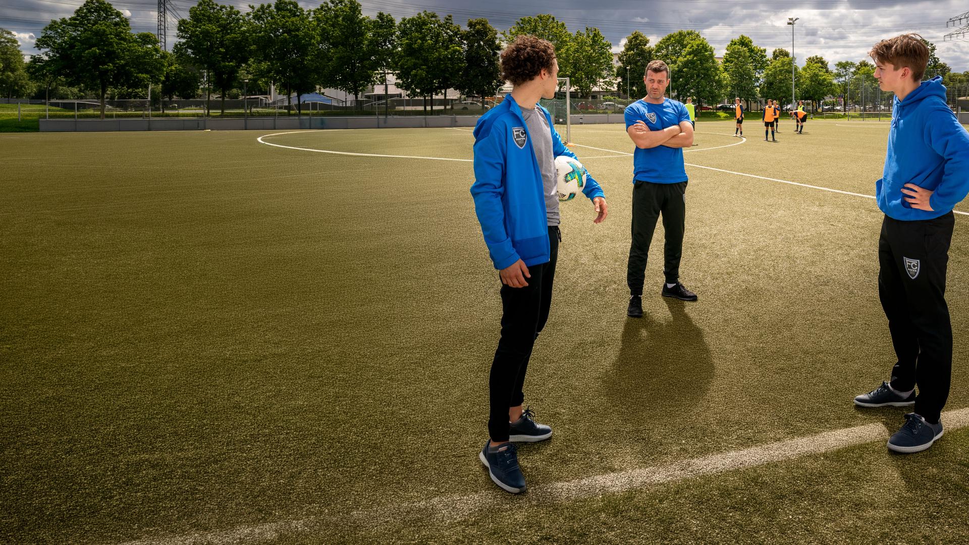 "Notruf Hafenkante - Fußball-Mütter": Noah Kruse (Jörg Rohde), Dominik Vogt (Riccardo Campione) und Lennard Kröger (Luke Matt Röntgen) stehen zusammen auf einem Fußballplatz. Dominik hat einen Fußball unter seinem linken Arm klemmen. Er blickt zu Noah, der links neben ihm steht. Noah hat die Arme vor dem Körper verschränkt und sieht Dominik an. Lennard steht Dominik gegenüber. Er hat die Arme in die Hüften gestemmt und sieht zu Noah.